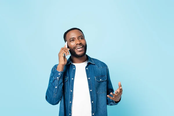 Усміхнений афроамериканський чоловік жестикулює і дивиться геть, розмовляючи на мобільному телефоні ізольовано на синьому — стокове фото