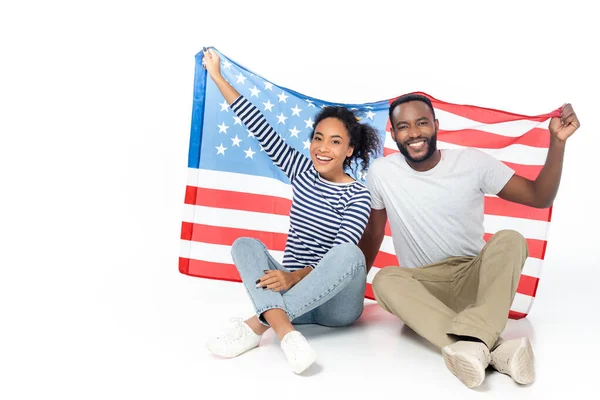 Joyeux couple afro-américain regardant la caméra tout en étant assis avec le drapeau des Etats-Unis sur blanc — Photo de stock