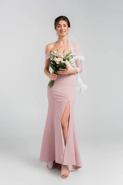 Ganzkörperansicht der lächelnden Verlobten in rosa Kleid posiert mit Strauß auf grau — Stockfoto