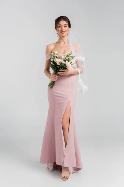 Full length view of smiling fiancee in pink dress posing with bouquet on grey — Stock Photo
