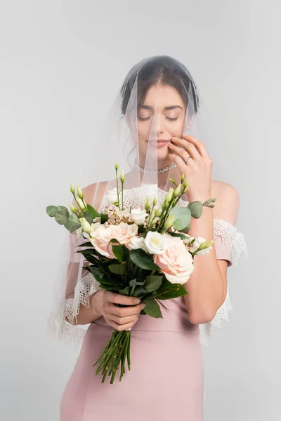 Junge Braut in Schleier hält Brautstrauß, während sie mit geschlossenen Augen isoliert auf grau steht — Stockfoto