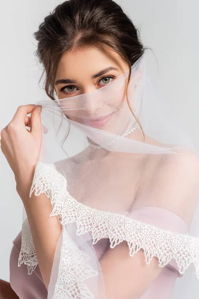 Coquettish fiancee covering face with veil while looking at camera isolated on grey — Stock Photo