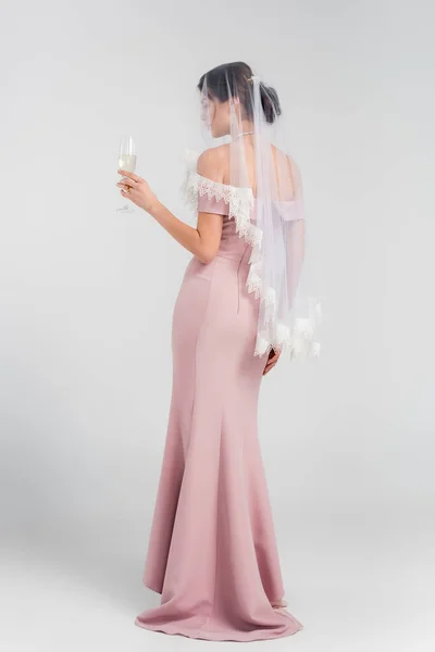 Ganzkörperansicht einer Frau in Brautkleid und Schleier mit einem Glas Champagner auf grau — Stockfoto