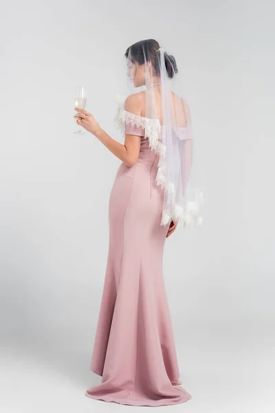 Full length view of woman in wedding dress and veil holding glass of champagne on grey — Stock Photo