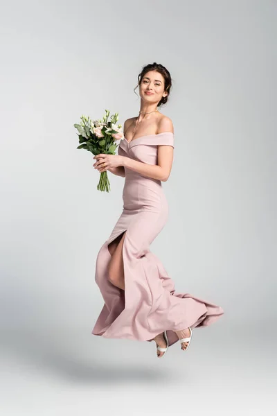 Full length view of happy bride holding wedding bouquet while jumping on grey — Stock Photo