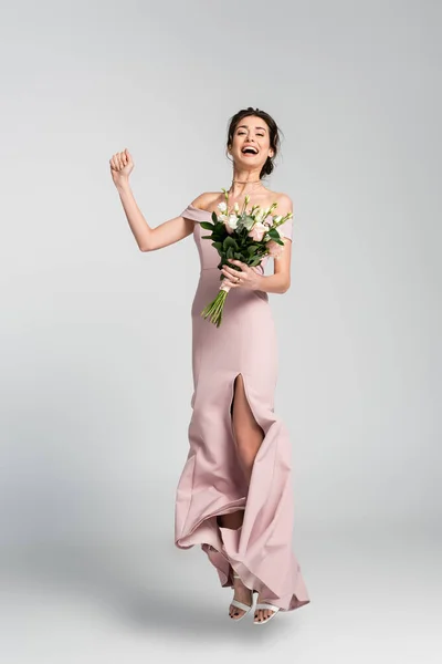 Full length view of excited fiancee holding wedding bouquet while jumping on grey — Stock Photo