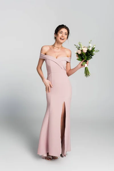 Full length view of excited woman in long wedding dress holding bouquet and looking away on grey — Stock Photo