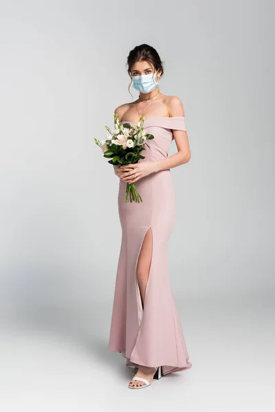 Ganzkörperansicht der Braut in medizinischer Maske posiert mit Brautstrauß auf grau — Stockfoto