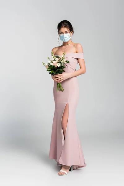 Full length view of bride in medical mask posing with wedding bouquet on grey — Stock Photo
