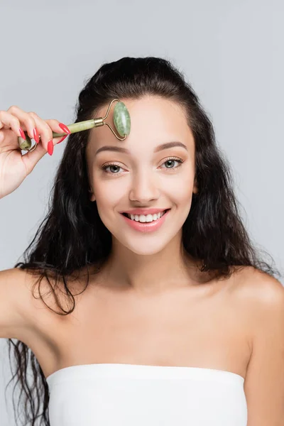 Happy young woman using jade roller while massaging face isolated on grey — Stock Photo