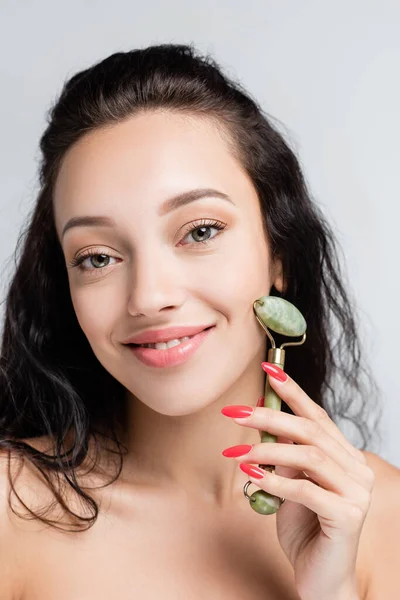 Smiling young woman using jade roller while massaging face isolated on grey — Stock Photo