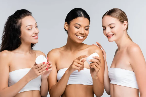 Alegres mujeres multiétnicas con hombros desnudos sosteniendo recipientes con crema cosmética aislada en gris - foto de stock