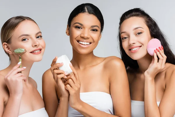 Mujeres multiétnicas sonriendo mientras sostiene el recipiente con crema cosmética, rodillo de jade y limpiador de silicona aislado en gris — Stock Photo