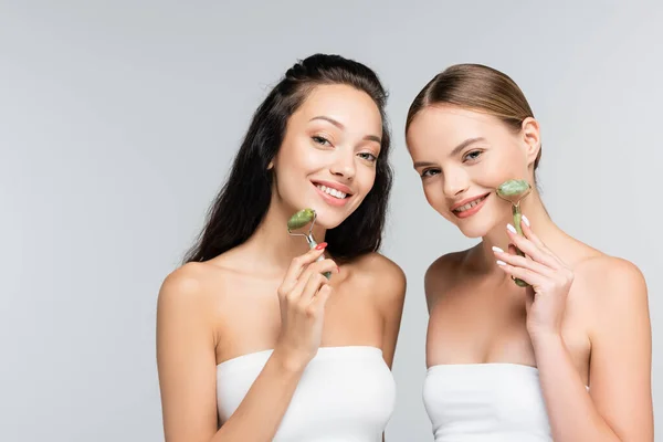 Mulheres alegres com ombros nus sorrindo enquanto segurando rolos de jade isolados em cinza — Fotografia de Stock