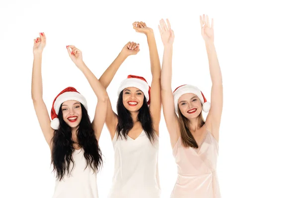 Mulheres inter-raciais alegres em chapéus de santa e vestidos de pé com as mãos acima das cabeças isoladas no branco — Fotografia de Stock