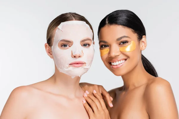 Mujer con mascarilla humectante cerca de feliz amigo afroamericano en parches de ojos dorados aislados en blanco - foto de stock