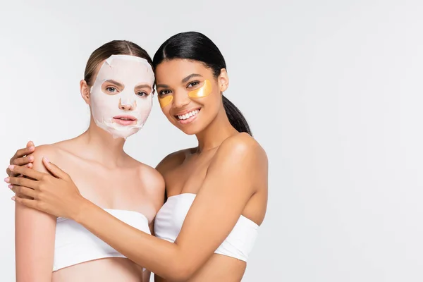 Mujer con mascarilla humectante cerca de alegre amigo afroamericano en parches para los ojos aislados en blanco - foto de stock