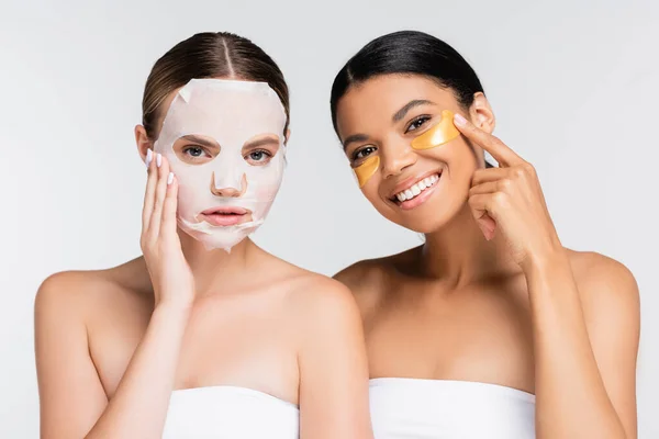 Mujer joven con mascarilla humectante cerca alegre amigo afroamericano en parches para los ojos aislados en blanco - foto de stock