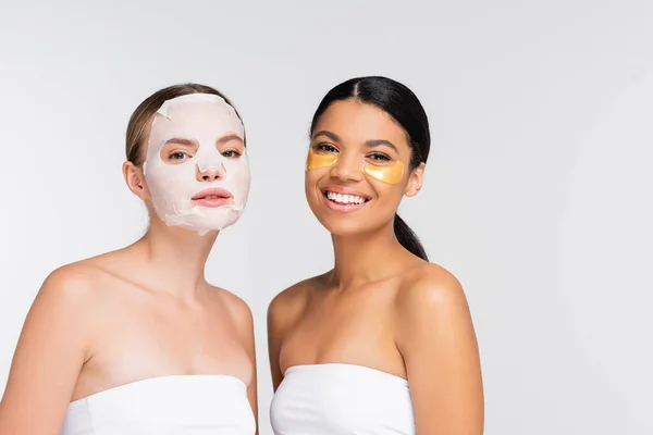 Mujer joven con mascarilla humectante cerca de feliz amigo afroamericano con parches en los ojos aislados en blanco - foto de stock