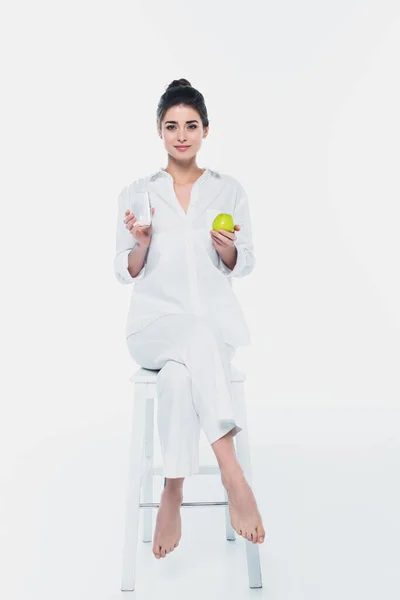 Mujer joven sosteniendo manzana y vaso de agua en silla sobre fondo blanco - foto de stock