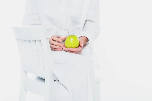 Обрізаний вид жінки в білому одязі, що тримає яблуко і склянку води на стільці ізольовані на білому — стокове фото