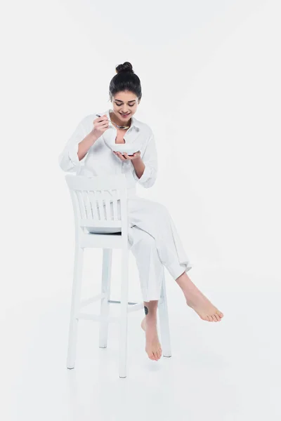 Усміхнена жінка тримає миску з крупами і ложкою на стільці на білому тлі — стокове фото