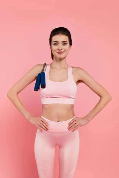 Sportiva sorridente con corda da salto e mani sui fianchi isolate su rosa — Foto stock