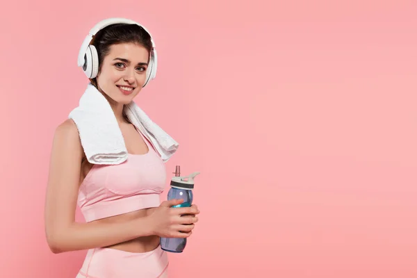Sportswoman smiling at camera while listening music in headphones and holding sports bottle isolated on pink — Stock Photo