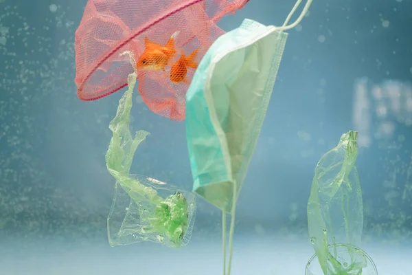 Red con peces de colores, máscara médica, bolsas de plástico y tazas en agua, concepto de ecología - foto de stock