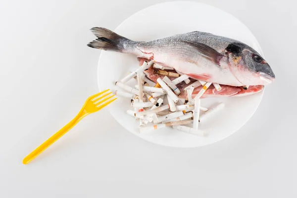 Вид сверху на пластиковую вилку рядом с тарелкой с выпотрошенной рыбой и концами сигарет на белом, экологическая концепция — стоковое фото