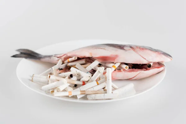 Teller mit Zigarettenenden und ausgenommenen Fischen auf weißer Oberfläche, ökologisches Konzept — Stockfoto