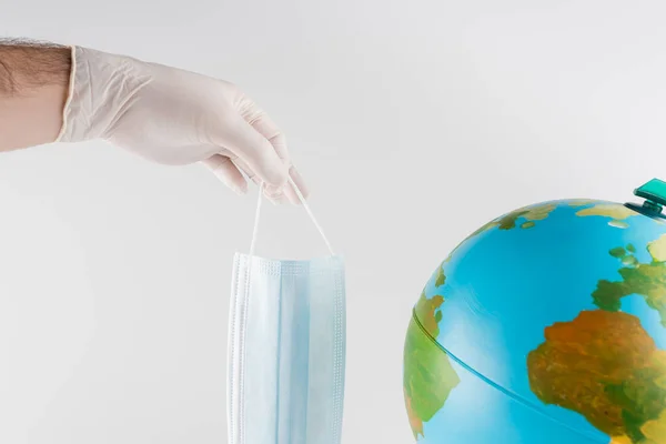 Vista recortada del hombre en guante de látex sosteniendo máscara médica cerca de globo aislado en gris, concepto de ecología - foto de stock