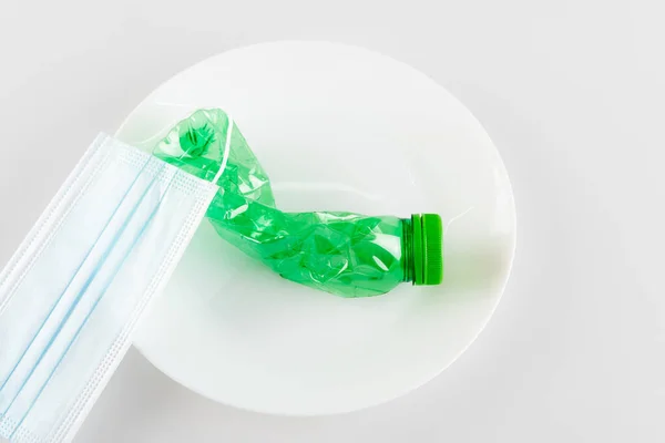 Вид сверху на медицинскую маску и смятую пластиковую бутылку на белом, экологическая концепция — стоковое фото