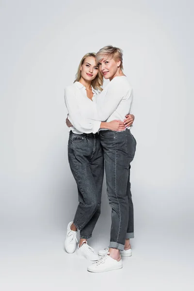 Full length view of stylish, joyful mother and daughter embracing on grey — Fotografia de Stock