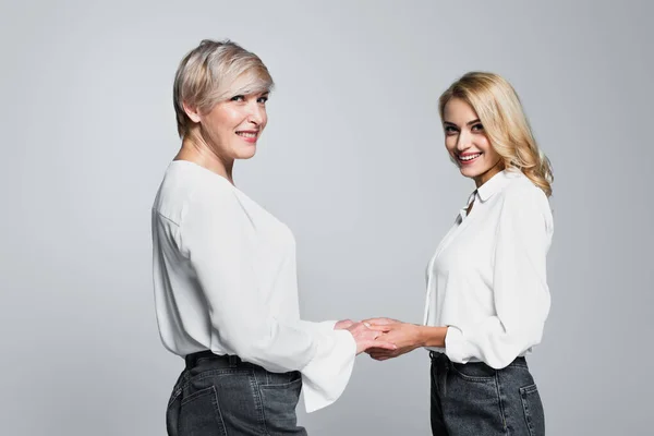Cheerful mother and daughter in white blouses holding hands isolated on grey — Foto stock