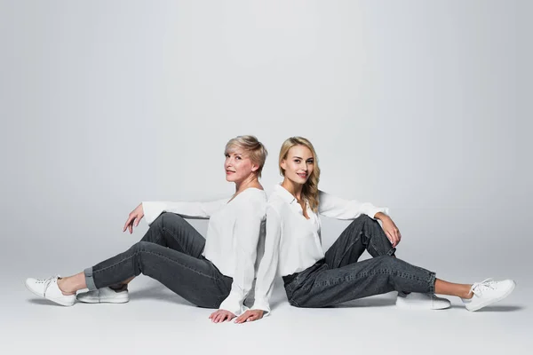 Full length view of mother and daughter in trendy clothes sitting back to back on grey — Fotografia de Stock