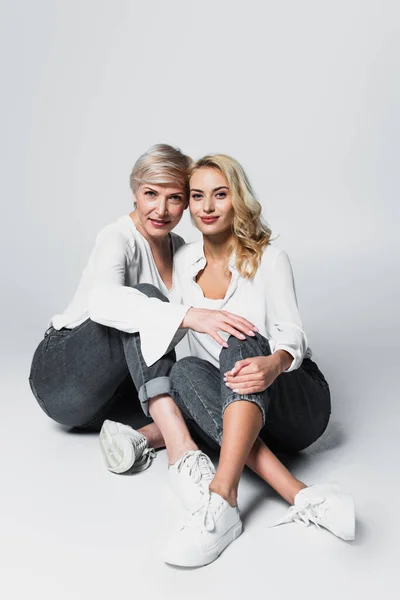 Full length view of mother and daughter in white blouses and jeans sitting on grey — Foto stock