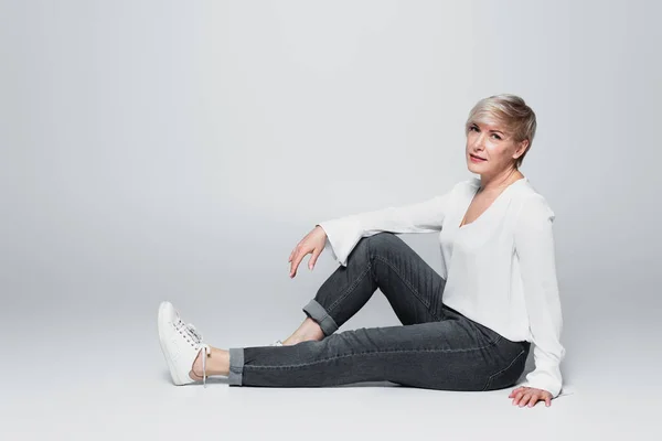 Full length view of stylish, mature woman looking at camera while sitting on grey — Fotografia de Stock