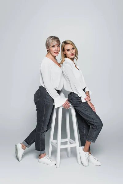 Вид в полный рост матери и дочери в джинсах и белых блузках, позирующих возле стульчика на сером фоне — стоковое фото