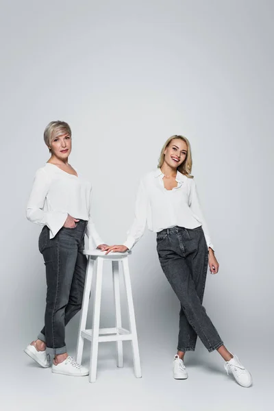 Full length view of mature woman with hand in pocket posing near high stool and young daughter on grey — Fotografia de Stock