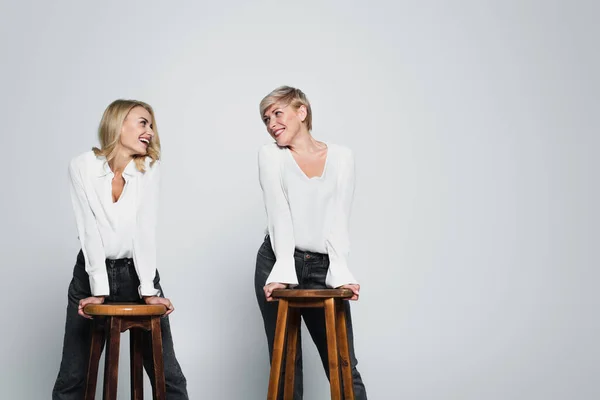 Joyful mother and daughter smiling at each other while posing near high chairs on grey — Fotografia de Stock