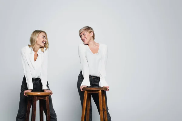 Joyful mother and daughter smiling at each other while posing near high chairs on grey — Foto stock
