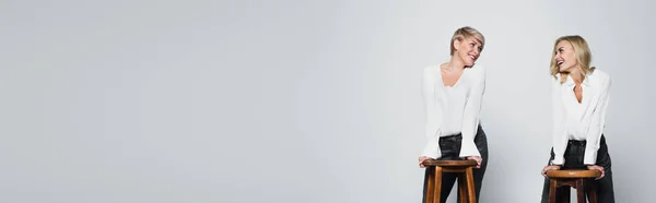 Cheerful mother and daughter looking at each other near high stools isolated on grey, banner — Fotografia de Stock