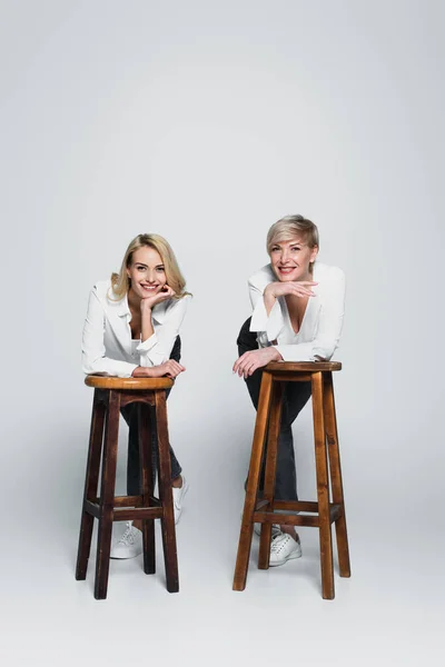 Full length view of cheerful, stylish mother and daughter looking at camera near high chairs on grey — Fotografia de Stock