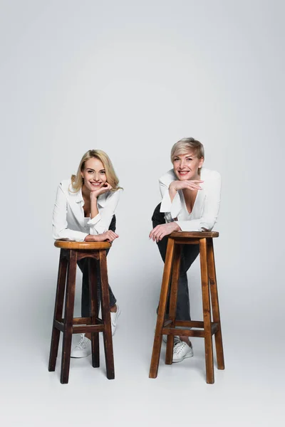 Full length view of cheerful, stylish mother and daughter looking at camera near high chairs on grey — Foto stock