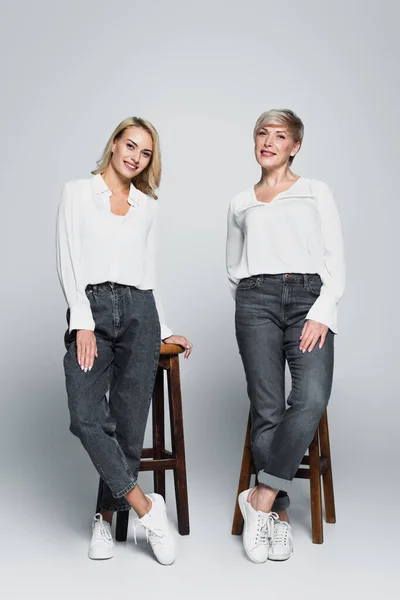 Full length view of smiling mother and daughter in trendy clothes posing near high stools on grey — Fotografia de Stock