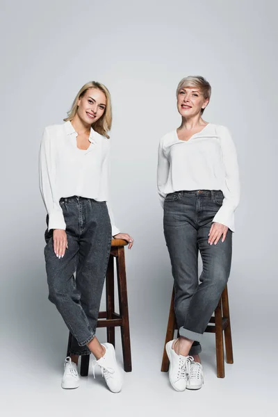 Full length view of smiling mother and daughter in trendy clothes posing near high stools on grey — Foto stock