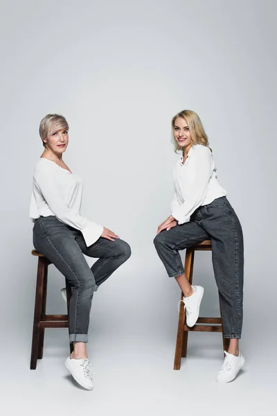 Full length view of mother and daughter in white blouses and jeans sitting on high stools on grey — Fotografia de Stock
