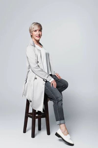Ash-blonde woman in trendy autumn outfit sitting on high stool on grey — Stock Photo