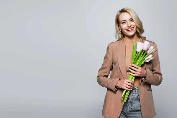 Joyful blonde woman holding fresh tulips while smiling at camera isolated on grey — Stock Photo